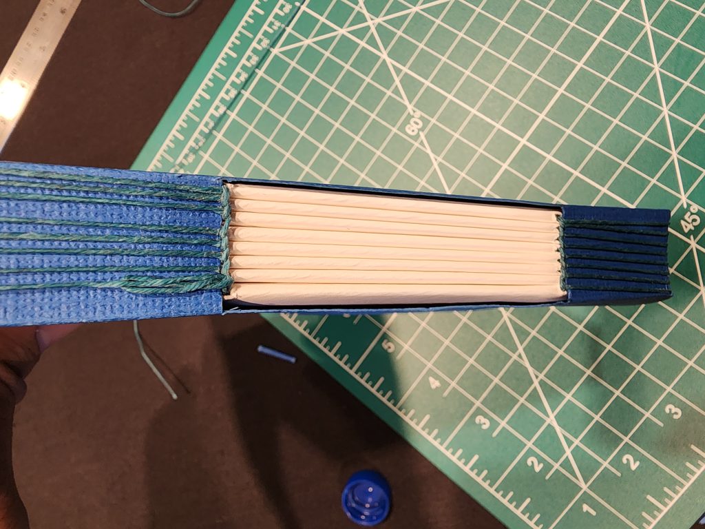 A cutting mat sits on a tabletop in the background, with some string and a ruler. The focus is on the spine of a book, the middle section of which is open to view the signatures behind it. The book cover is blue, and a blue/green thread is seen binding the signatures to the cover. A loose stitch causes a small loop of thread to be seen on one signature.