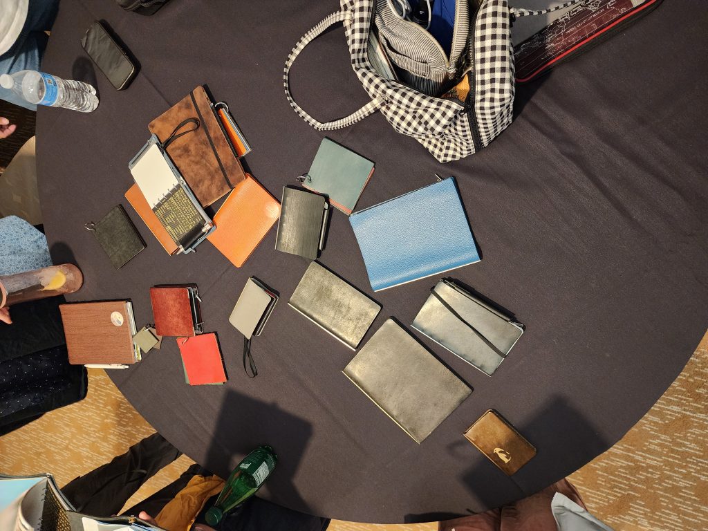 A circular table covered in a black cloth, covered with 16 or more notebooks and an open bag. The notebooks are from Plotter, made of leather, and in a variety of colours. Each is well used and filled with various papers.