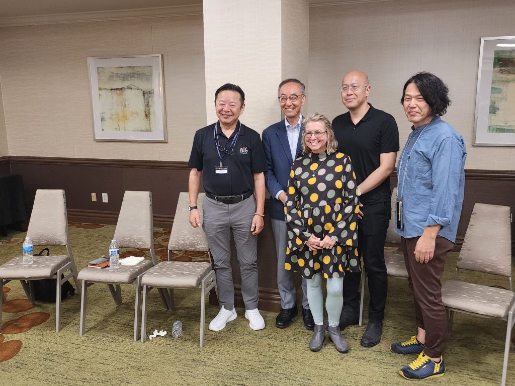 Five people stand smiling, lined up at the front of a room. Several chairs around them, and a couple modern abstract paintings on the wall behind. Four men of roughly equal height, left to right, Yukio Nagahara of Nib Shaper, Naoyuki Takeuchi of Nagasawa Stationery, Daryl Lim of Musubi, and Sajori Takahashi of Sey. In front of them stands Lisa Vanness of Vanness Pen Shop, a head shorter than the rest. They pose for pictures from the audience after a panel discussion.