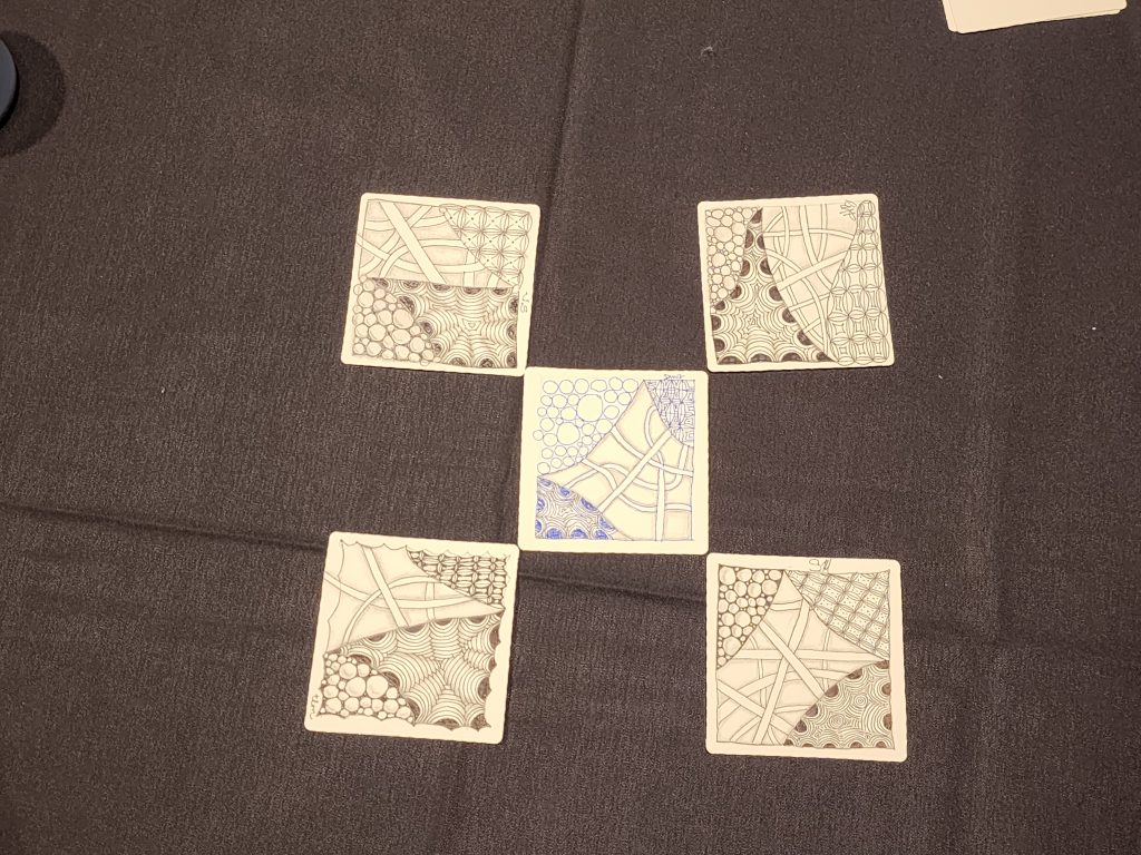 A black tablecloth with 5 square paper sheets spread in a checker pattern on top. Each paper square was done by a different student, showing their own creative work, and is decorated with 4 different patterned sketches. Crescent Moon, Hollibaugh (overlapping bridges), Bales (decorated grids), and Tipple (bubbles).