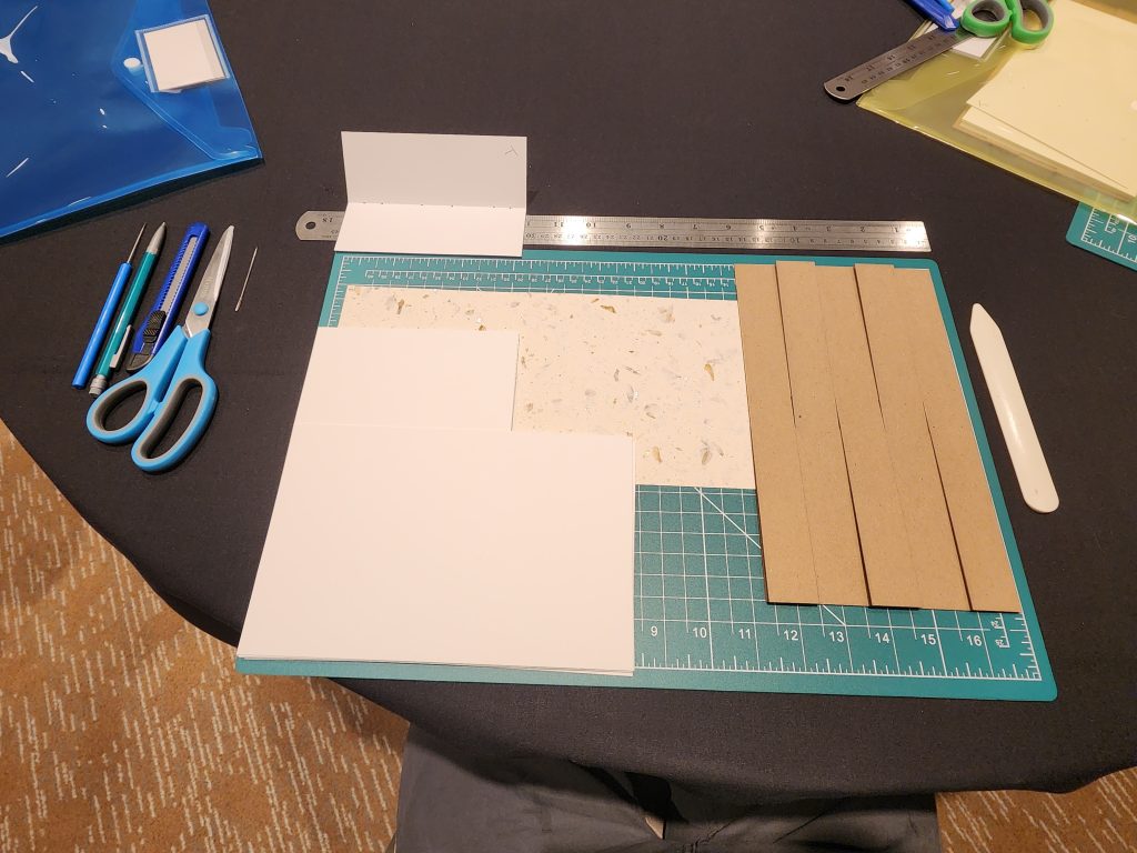A circular table with bookbinders tools laid out on top. A cutting mat, ruler, cover template, hand-pressed cotton and flower paper for the cover, a bone folder, scissors, sewing needle, awl, box cutter and mechanical pencil.