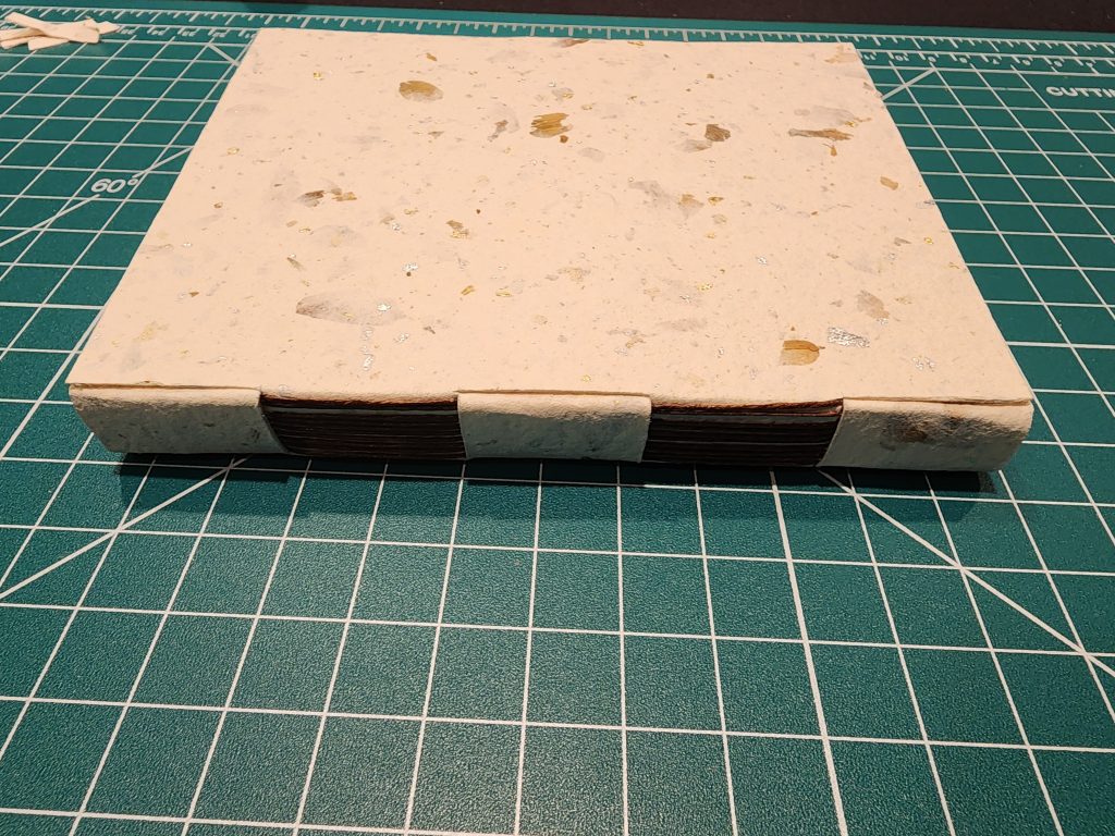 A cross structure bound book rests on a cutting mat, showing the spine and threadwork, along with the back cover. The cover is made of a hand-pressed, textured cotton paper, with bits of flower petal and gold flecks throughout.
