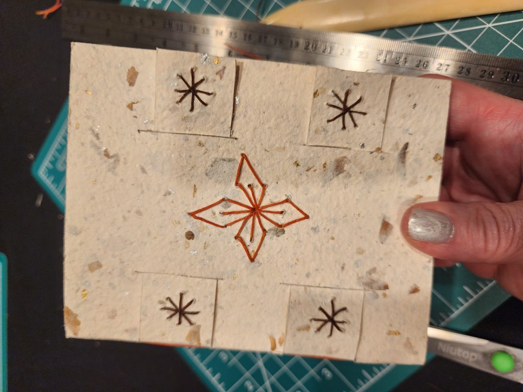 An image focusing on a small, A6 sized, book, with a hand pressed flower paper, white cotton with flecks of flower petals and gold specks seen throughout. On the front is embroidered a 4 petal flower with red string, and 4 asterisks in brown string.