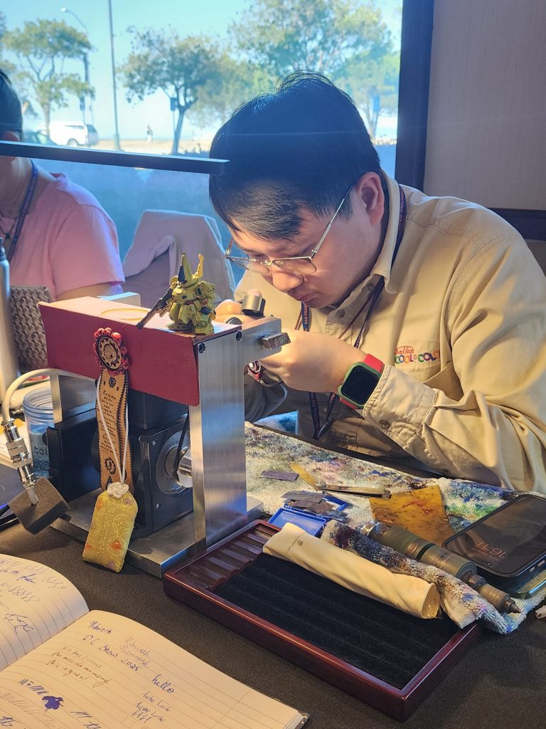 Matthew Chen, of Matthews Nibworks, a nib grinder, sits in front of his table, with various tools (lathe, lights, mesh, etc) in front of him. He focuses on a pen in hand, inspecting the nib with a jewlers magnification loupe.