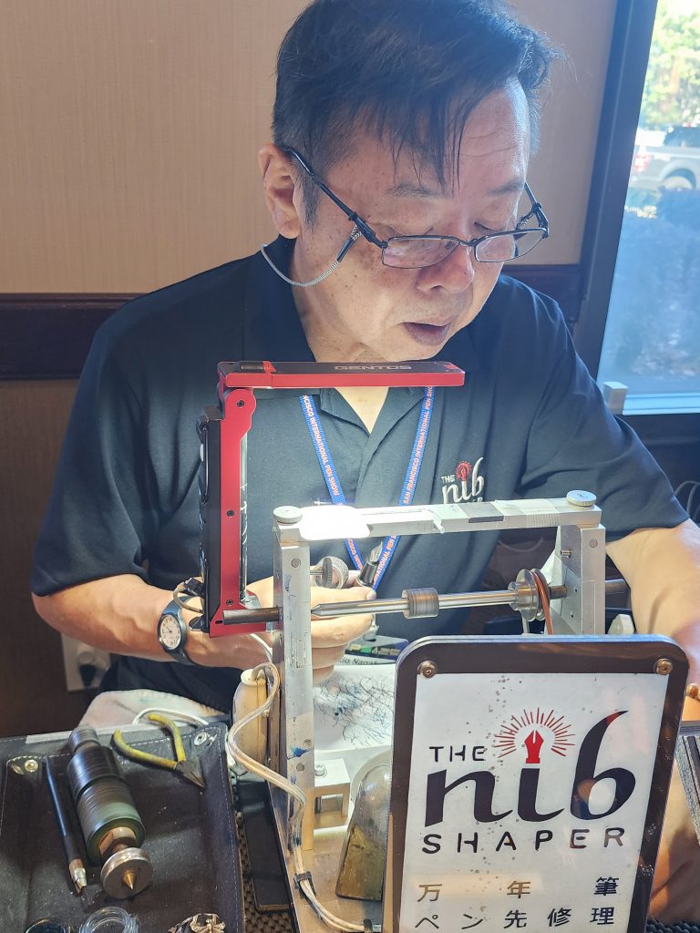 Yukio Nagahara, the Nib Shaper and expert nib meister, sits in front of his table, with various tools (lathe, lights, mesh, etc) in front of him. He focuses on a pen in hand, beginning his work to shape the nib.