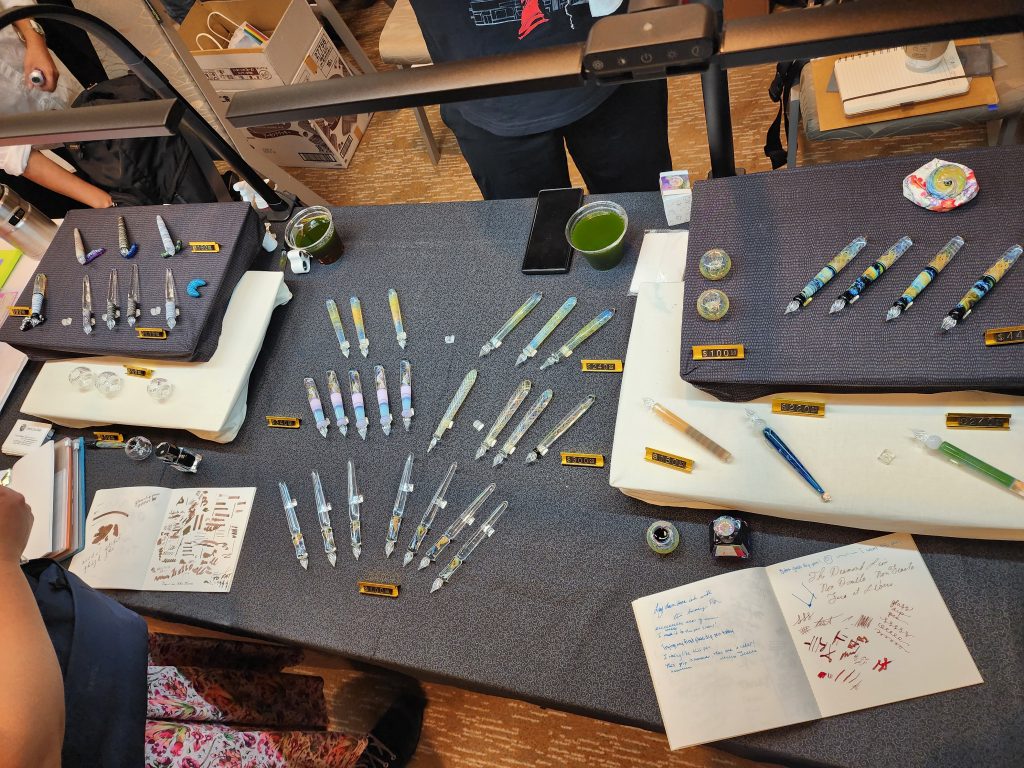A table covered in grey cloth, with several raised tiers. Glass dip pens are arranged in a display all over the table, along with glass pen holders, several small glass caterpillar pen rests, and a couple open notebooks and ink bottles for customers to test pens. The pens range in shape and colours, from tapered handles to traditional cigar shapes, with grip sections made of spheres or cylinders.