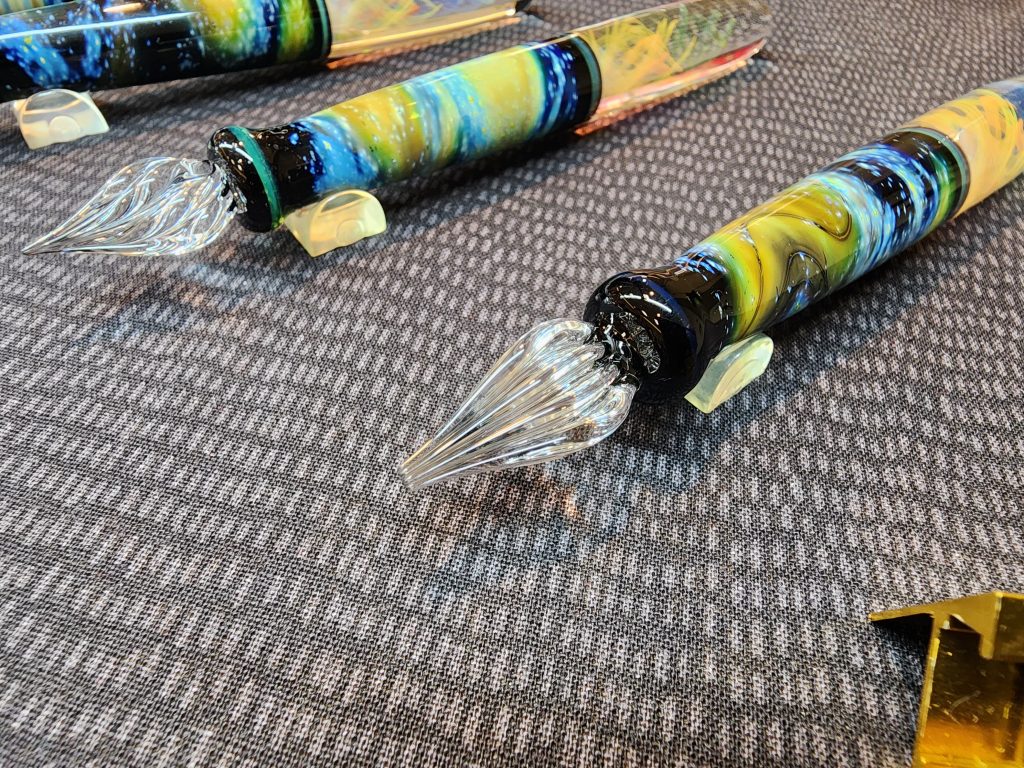 A closeup image of a couple glass dip pens. The colourway on each pen is a swirled mix of blue, green, ochre, black and various others, lending to its namesake, "Cosmic Dip Pen". The pen in the foreground has a wide glass dip nib.