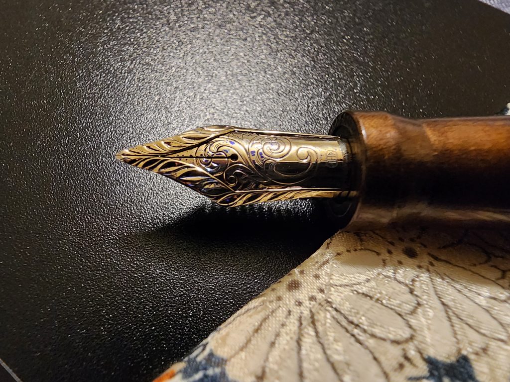 A closeup photo of a beautifully crafted stacked, gold nib, with an intricate design. Some small ink spots can be seen on the nib, it rests on a black background with part of a floral pen sleeve underneath the pen.