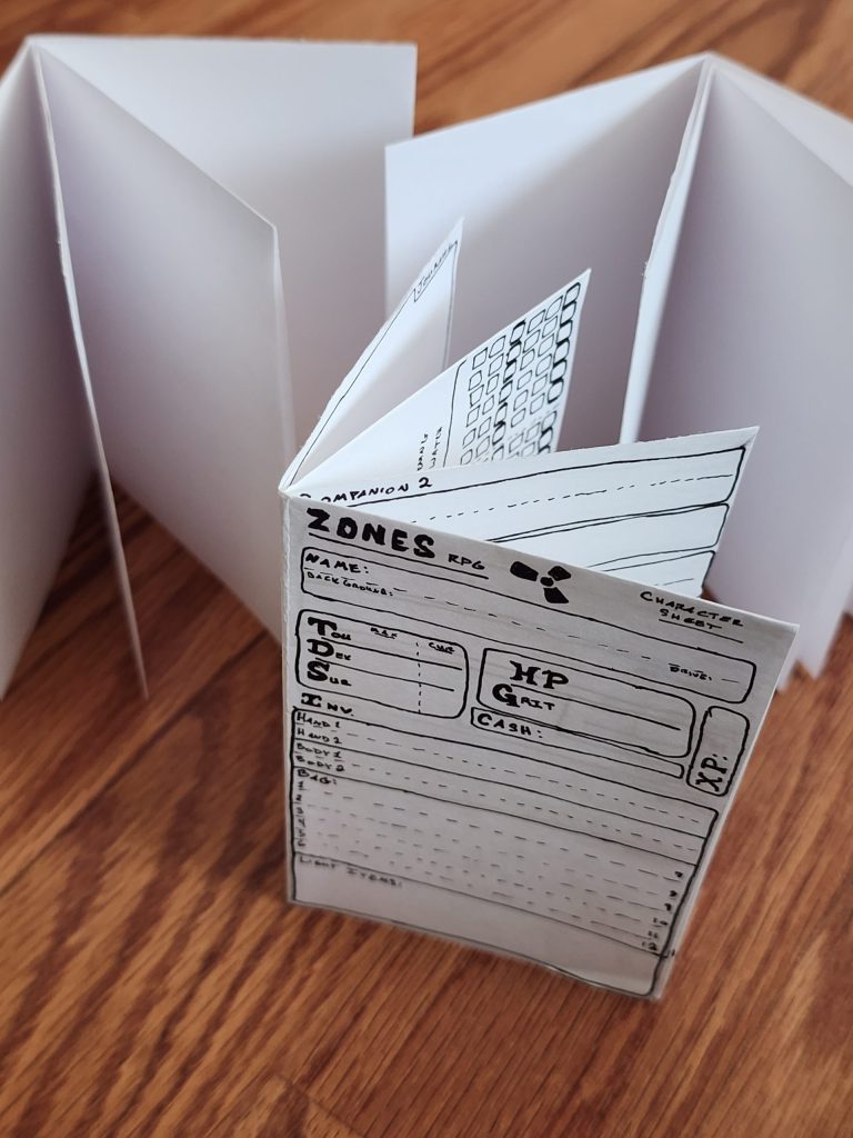 Three sheets of paper, folded into 8-page mini-zines, stand on end, pages splayed open. One of them has been turned into a mini character sheet for the Zones RPG.