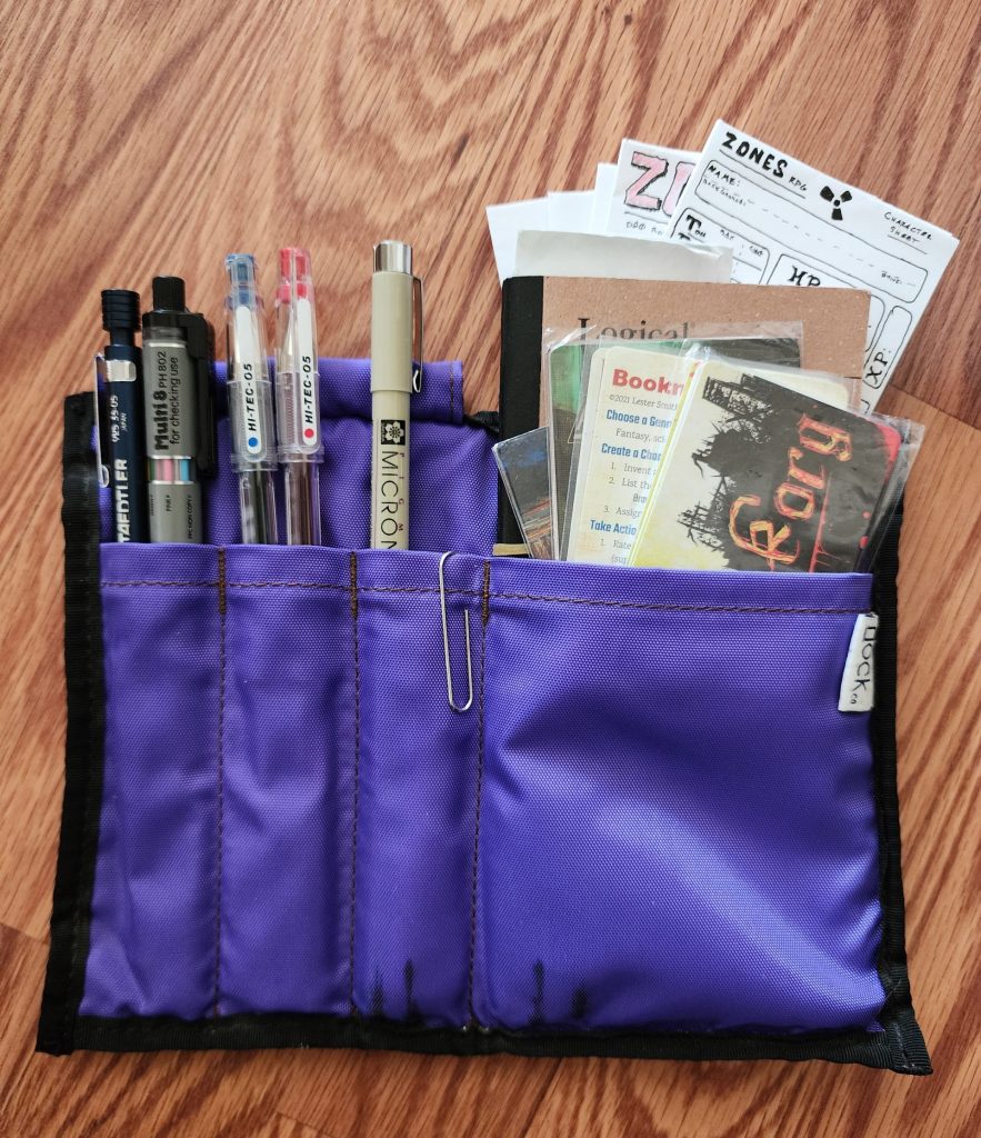 The Nock Co Hightower, a bifold case constructed with a 1000D nylon material with black border trim. The case is open, exposing an amethyst purple interior, 3 pen pockets on the left, a deep notebook pocket on the right. The contents of the case are exposed, showing how much can fit within.