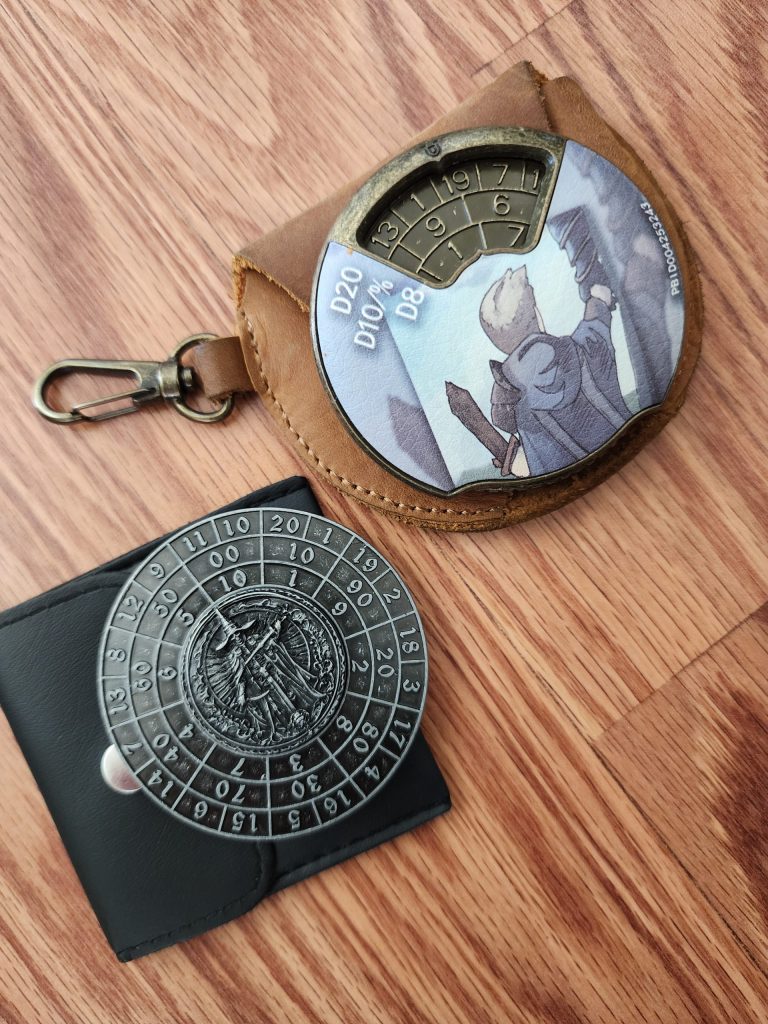 Two metal dice spinners, comprised of concentric rings filled with numbers representing various polyhedral dice (d4, d6, d8, d10, d12, d20). They are two different designs, one a spinning disc enclosed in a metal shroud, the other a central bearing with rotating outer ring. They both sit on top of small carry pouches, to protect them.