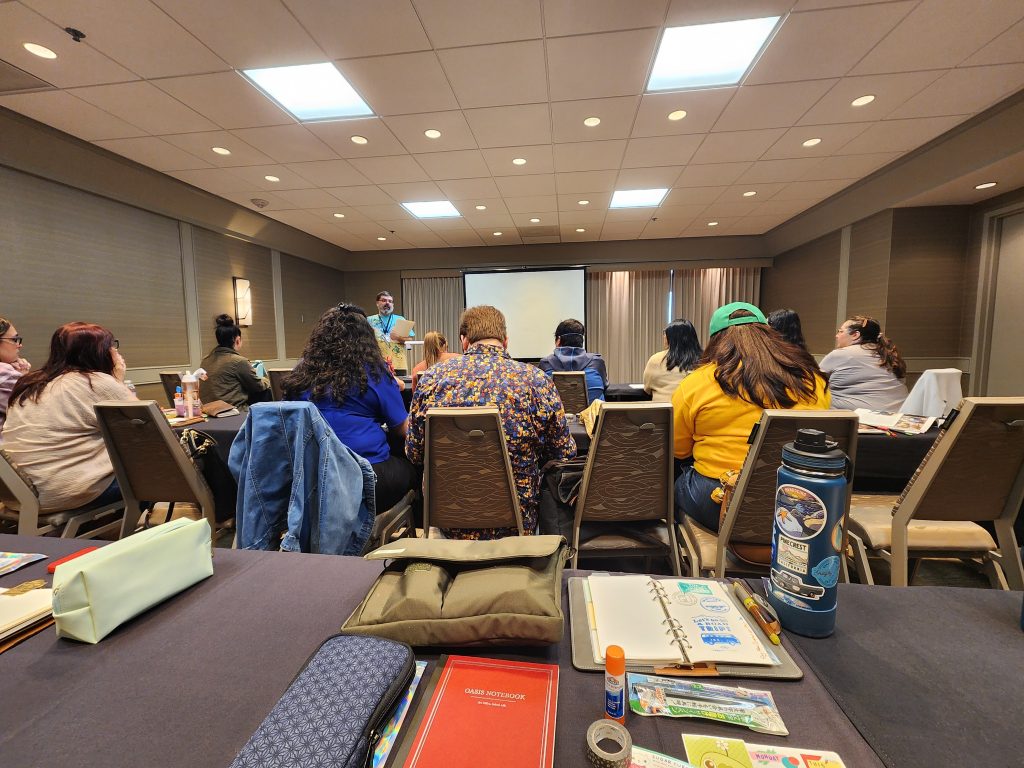 A variety of stationery items - pouch, notebooks, tape, glue, stickers, etc - sit on a table in a classroom style setting. A teacher stands in front, addressing all the people sitting at the tables, ready to learn about Creative Journaling.