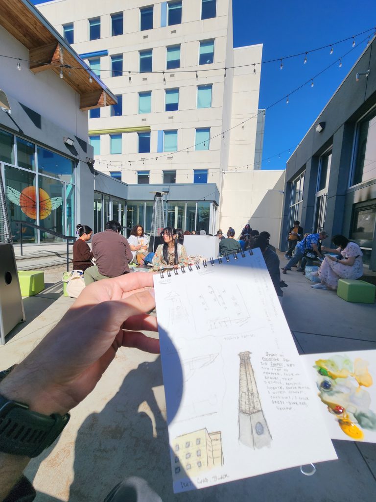 Sitting in an outdoor courtyard, surrounded on 3 sides by buildings, a disembodied hand holds up a small paper pad, with artistic scrawlings at an urban sketching class. In the background a large group of people sit, similarly focused on their own practice.