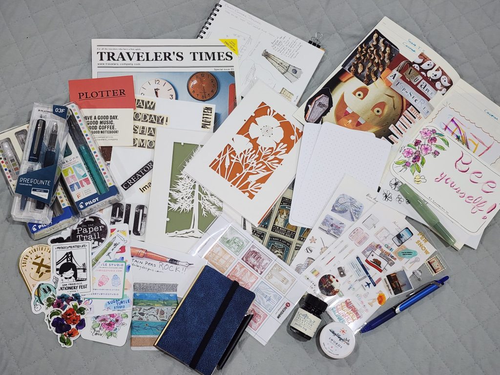 Sitting on a grey blanket is a collected pile of stationery and ephemera. Notebooks, notecards, laser-cut greeting cards, a couple fountain pens, a blue PLOTTER notebook cover, a bottle of ink, a roll of washi tape, and a bunch of stickers.