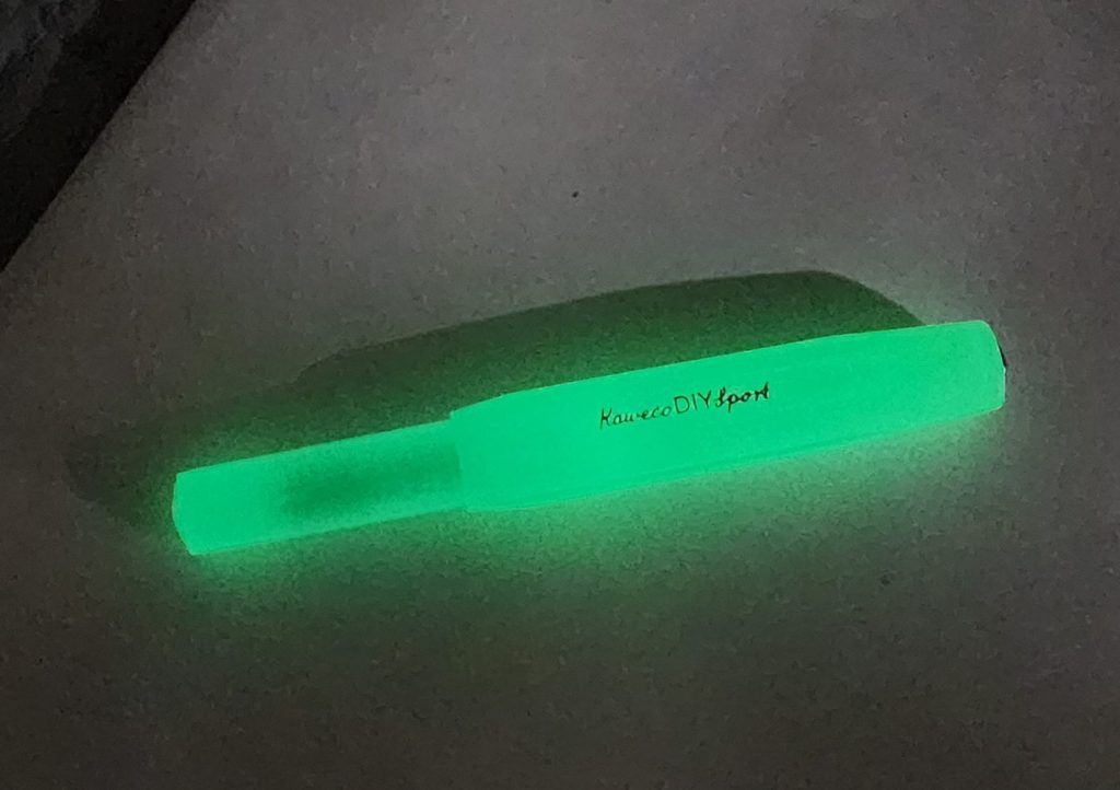 A grainy image of a Kaweco DIY Sport fountain pen, taken in a dark room. The fountain pen glows in the dark, emitting a bright green glow. The etched name "Kaweco DIY Sport" visible in the pen cap.