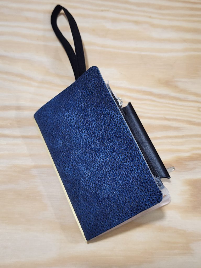 A small, blue notebook cover, with a bumpy texture sits on a wooden tabletop. The PLOTTER Kurozan Mini5. An elastic band sticks out of the top and rests on the wood table, with a narrow pen holder sticking out of the right side of the cover.