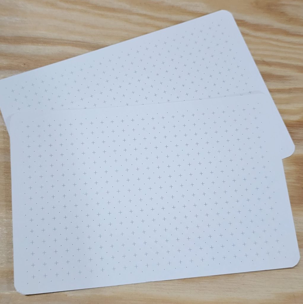 A pair of white 4x6 notecards, covered in light-grey printed alternating dots and crosses sit atop a wooden table.