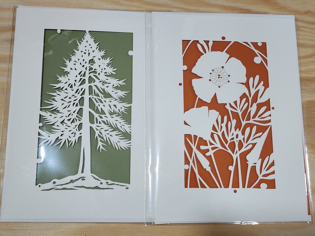 A pair of laser-cut greeting cards sit on top of a wooden table. On the left, the outline of a pine tree is cut from the card, with a green paper insert behind it making the cutout stand out. On the right, a set of poppy flowers with small stems are cut out, with a deep orange sheet behind. Both cards are still in their cellophane wrappers.