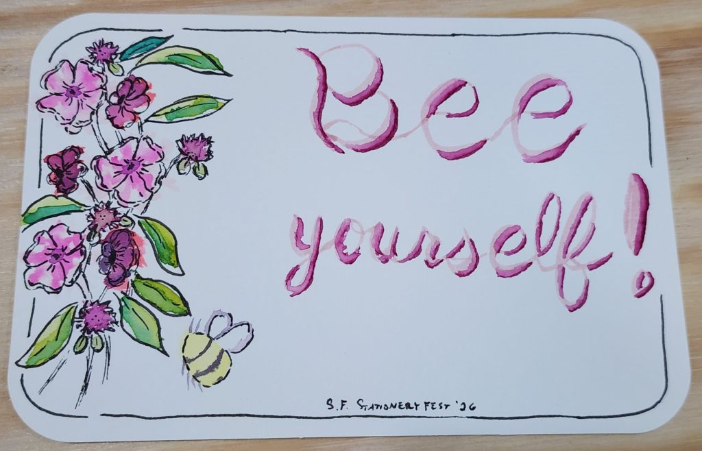 A white postcard with rounded corners sits on a wooden table. The postcard is watercolour paper and has several pink and purple flowers on the left, with leaves in a few shades of green. A large bumble bee has been painted in floating near the flowers. On the right, the text "Bee Yourself!" has been painted as well.