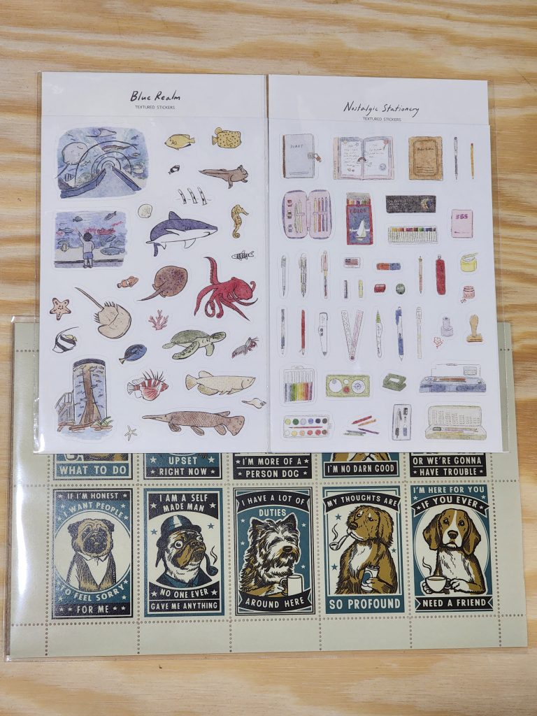 A trio of sticker packs sit on a wooden table. One has a variety of aquarium and aquatic creature stickers. Another has various stationery items, such as pens, markers, and journals. The last one is a series of gentlemanly dogs, with various bits of clothing, drinks and pipes. Each dog sticker has a different statement around it, such as "My thoughts are so profound".