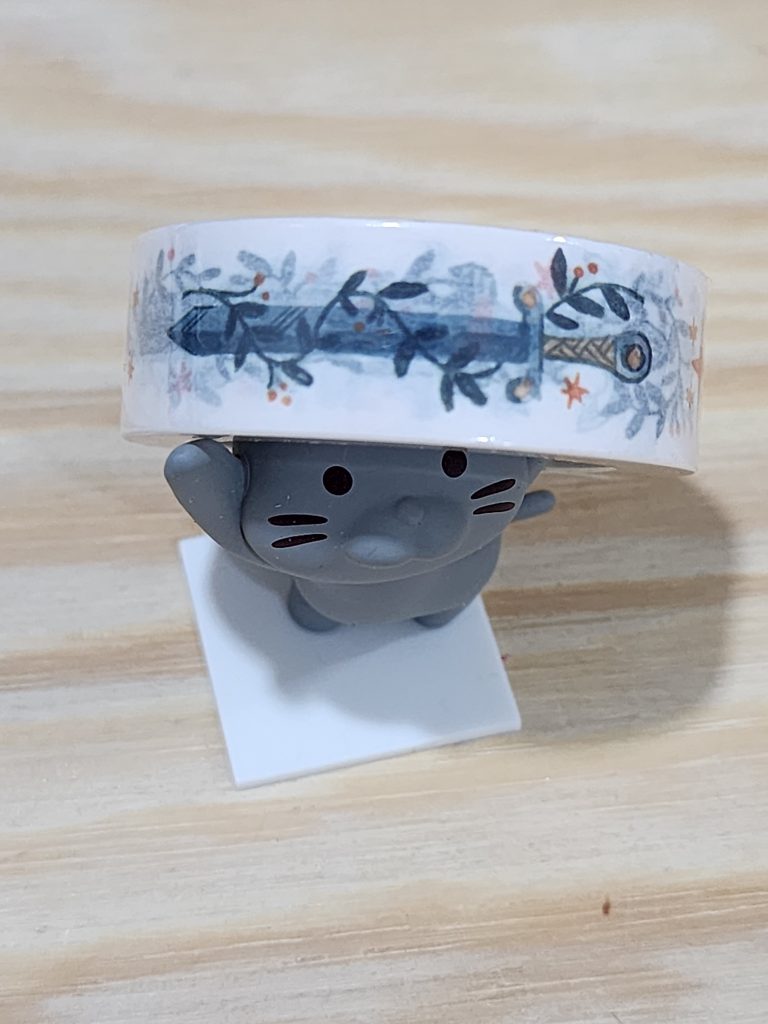 A small, grey, cartoonish cat stands with its front legs in the air to make a pen stand. Instead of a pen, the cat is holding up a roll of washi tape, themed with various fantasy swords.