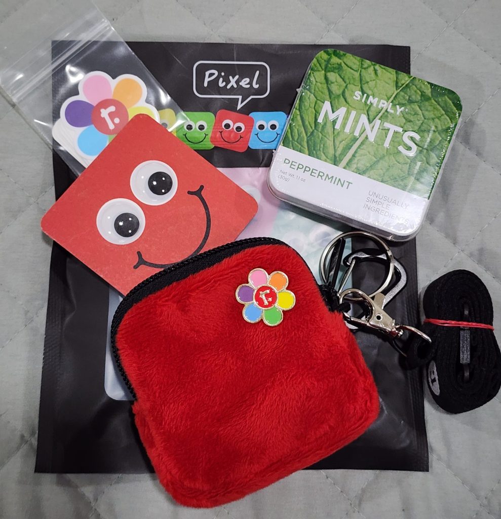 On top of a bag labelled "Pixel" sits a small, red, fuzzy bag. A multi-coloured pin is attached, and spilling from its opened pouch is a red cardboard square with googly-eyes and a big smile, a pack of small stickers, and a square mint tin.