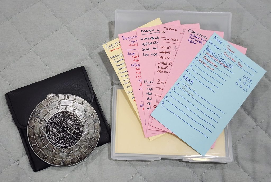 Two items sit on a grey blanket. A small dice spinner, comprised of a rotating exterior dial and stationary inner ring to imitate the results of random dice rolling. A plastic card case, the lid open, sitting on top are several cards in yellow, red and blue. Each card has small, handwritten text, taking notes on a solo rpg game.