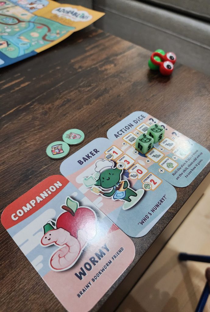 An image showing part of a tabletop, with a game of Adorablins in session. Three cards sit in a row, the Baker character, Worm companion, and an Action Dice card. Two dice, showing 6's, sit on the action dice card, above the row of cards are 2 "snack" chits. The edge of a paper map is visible in the corner of the image. A set of googly eyes sits near the edge of the table.
