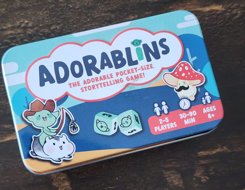 The top of a small game tin for the game Adorablins. The imagery on the lid shows whimsical art of a small goblin smiling, wearing a cowboy hat, and riding a hamster. A pair of dice with small goblin faces. A mushroom with a monocle and a moustache. It indicates the game is meant for 2-5 players, runs for 30-90 minuets, and is meant for ages 8+. "Adorablins. The adorable pocket-size storytelling game!"