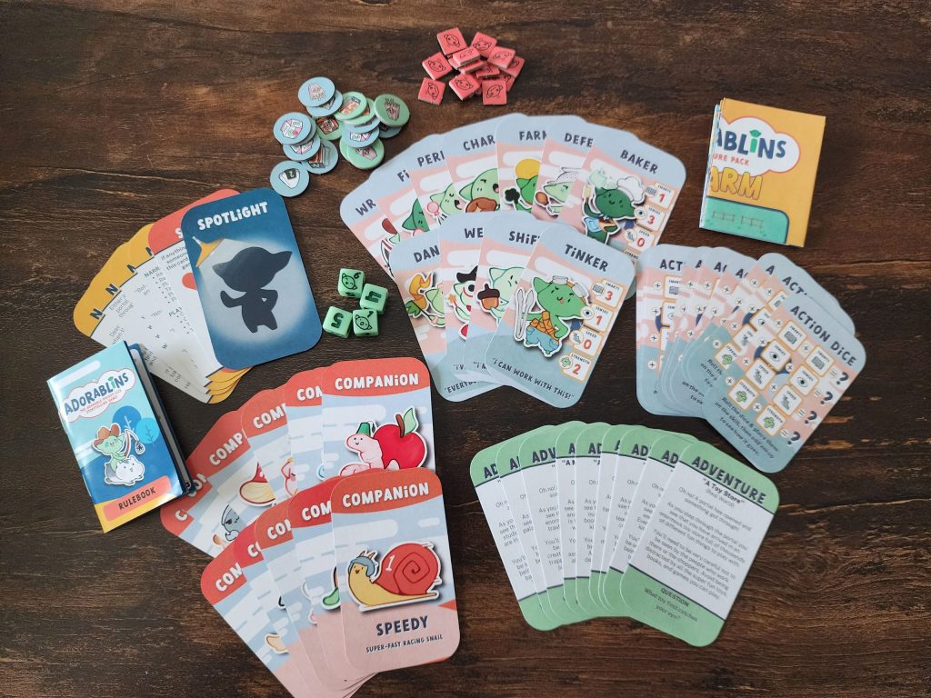 A tabletop with a variety of cards from the Adorablins game, fanned out, two piles of small chits, a folded paper map, a small rulebook, and a set of four six-sided dice. There are Character cards, each with an image of an adorable goblin with a specific job - Baker, Tinker, Defender, Farmer, etc. Companion cards depicting goblin companions, such as Speedy the racing snail, Wormy the worm, and more. Adventure cards, which detail some pre-made adventures for the Adorablins. A set of Narrator cards, including a spotlight card and an X card. Chits depict a frazzled goblin and a variety of snacks. Action Dice cards indicate how to roll the dice and add them together to determine a result.