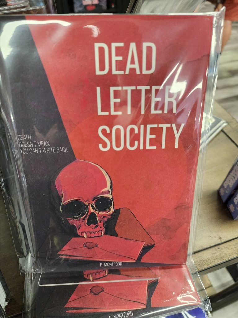 On a store shelf sits a zine, the cover red with a black streak down one side. It depicts a redskull sitting atop a journal and envelope. The title reads, "Dead Letter Society"