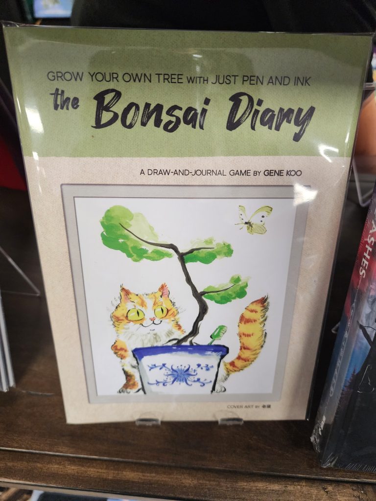 On a store shelf sits a zine, its cover part green part tan, it reads, "The Bonsai Diary, grow your own tree with just pen and ink". An image of a bonsai tree growing in a fanciful glass or stoneware pot, with a cat watching a butterfly.