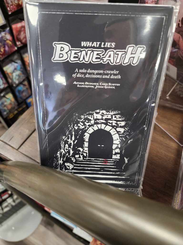 On a store shelf sits a zine, black with white text reading, "What Lies Beneath. A solo dungeon-crawler of dice, decisions, and death." A simple image of stark white against the black background depicts stairs leading up to a doorway, with a set of white eyes staring down the stairs.