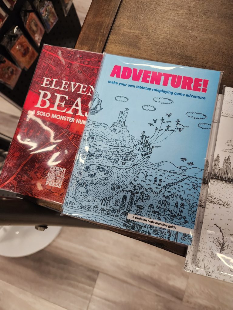 On a store shelf sits two solo-focused TTRPG zines. Adventure, a blue cover with bright pink header font, and simple graphics depicting a town with its center on a floating island. Also, Eleventh Beast, a solo monster hunting game, the cover is red, depicting an overhead view of the map of a city.