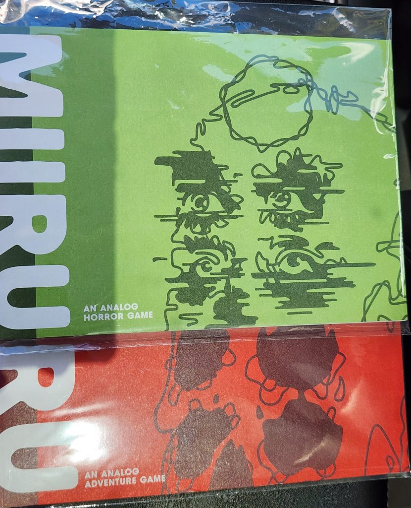 Two bookletes, one sitting atop the other. The top one is green, with darker green lines making an image of 2 sets of eyes on a face, white text reads "MIIRU" (Miru 2). The bottom one is red, with what looks like part of a skull in darker red, also with 2 sets of eyesockets. The white text on its cover reads MIRU (Miru 1). Both are solo focused analog games.