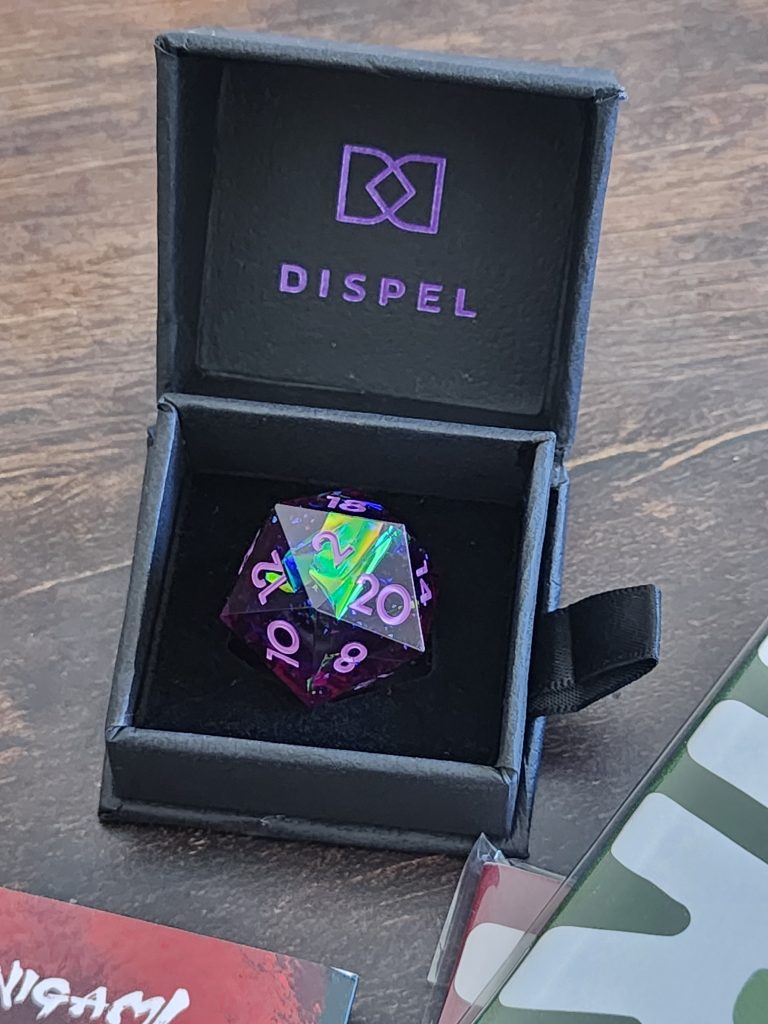 A small black box sits propped open, holding a single twenty-sided die. The interior lid reads "Dispel". The die sits on a small foam block, its overall colour ranging from ping to purple, with a shining glimmer of blue/green inside. The numbers are painted in pink.