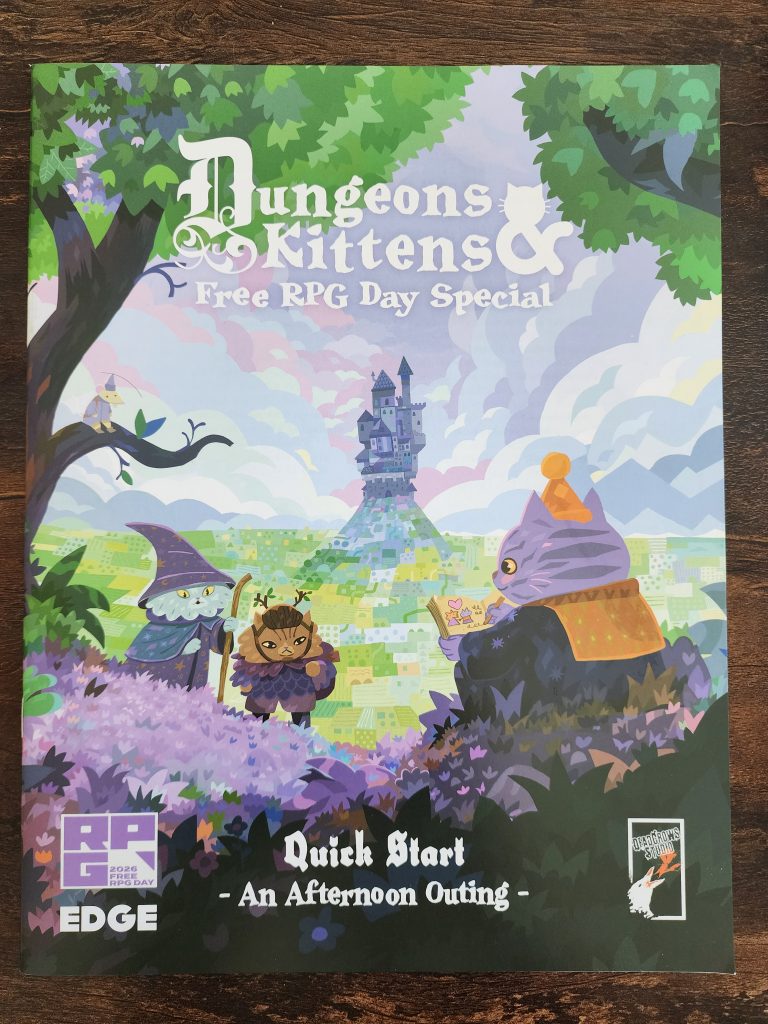 On a tabletop sits a booklet, the title reads "Dungeons & Kittens Free RPG Day Special, An Afternoon Outing". The cover depicts 3 cats, one sitting and writing in a book, two travelling up a hill to meet the third. In the background is a sea of sprawling green farmland, and a tall castle atop a hill.