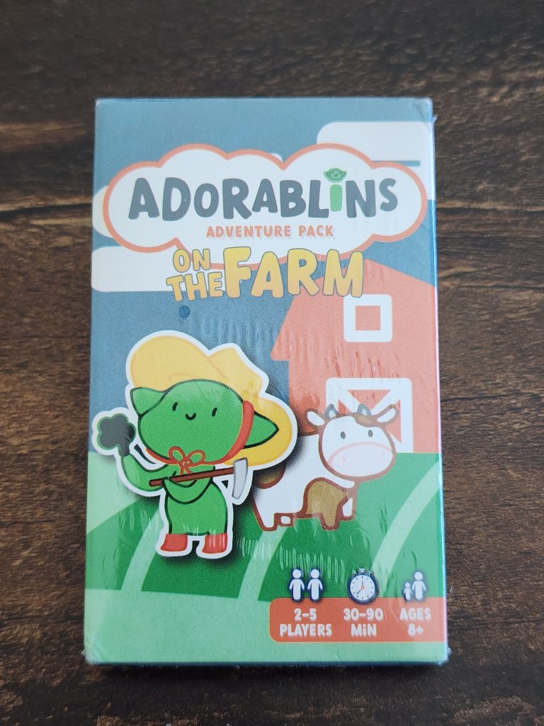 On a table sits a small cardboard box reading, "Adorablins Adventure Pack, On the Farm". An adorable goblin character stands in front of a cow and a barn, holding brocolli and a hoe.