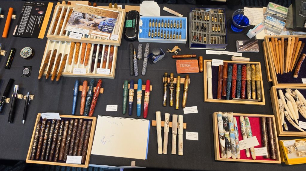 A table from the SF Pen Show, covered in a variety of pens, holders and nibs. Some are wood, some made of antler, some urushi laquered, and some with makie artwork.