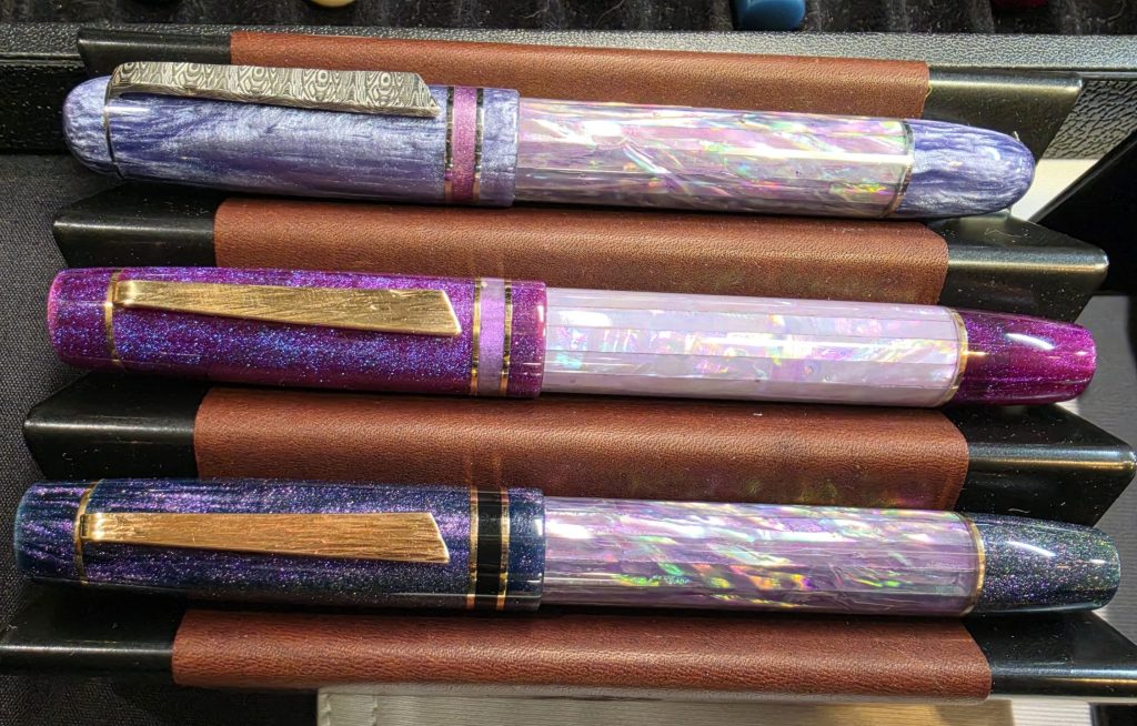 A set of 3 fountain pens. Sparkly, shiny, with various shades of purples and blacks. The body of each pen is faceted abalone. The clips made of worked metal.