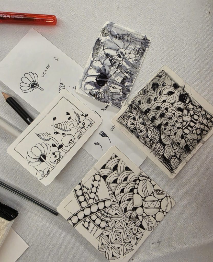 Several paperstock cards sit on a white table sheet, a few pens and pencils scattered around. Two of the paper cards have "zentangling" artwork, a series of geometric shapes, circles, and arches making patterns. Another two cards have make-believe plants and organic sketches, looking similar to flowers, mushrooms and leaves.