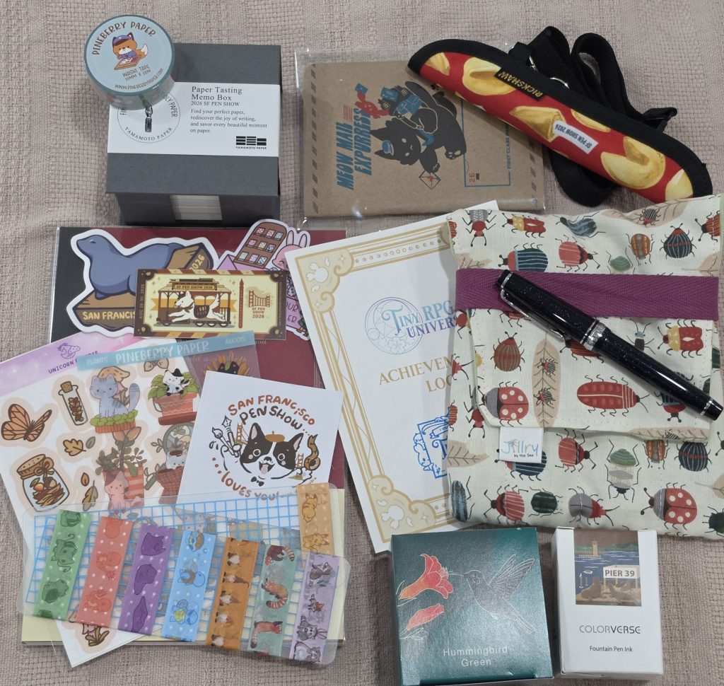 An orderly collection of stationery items lay on a blanket, showcasing a small assortment of goods collected from the SF Pen Show. A notebook, sticker sheets, die cut stickers, a pocket notebook, pen sleeve, pen case, fountain pen, fountain pen ink bottles, washi tape roll, and a small square box of paper labelled "Paper Tasting Memo Box" from Yamamoto paper.
