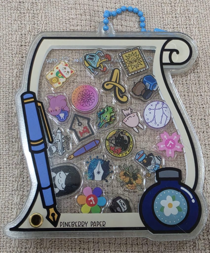 A flat, acrylic keychain ornament, known as a Shaker Keychain, in the shape of a scroll of paper with a pen and ink bottle, lays on a blanket. The charm is two pieces of acrylic that slide apart. Inside the ornament are over a dozen small acrylic charms, depicting various stationery vendor logos and artworks. The charms "shake" inside the ornament.