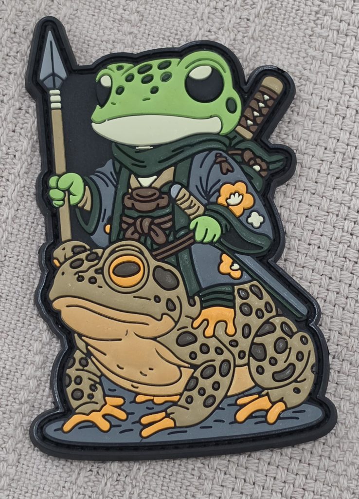 A 3 inch hook-and-loop patch, depicting a cute, green frog warrior. Its wearing what looks like a traditional Japanese Kimono and Hakama. It wields a spear, wears a sword, and is riding the back of a brown toad.
