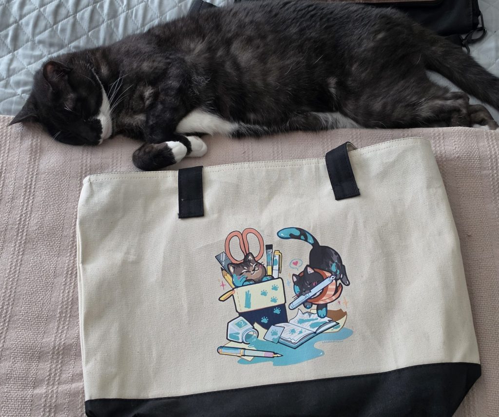 A cat lays on a bed, sprawled out next to a canvas bag that depicts two cats making a mess of stationery items (pens, ink, notebooks, etc).