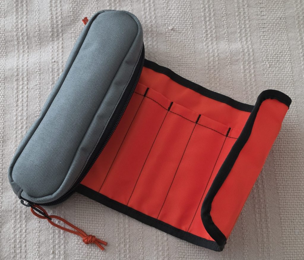 A Rickshaw Bags pen roll, a re-release of the Nock Co Brasstown. A canvas bag in grey, with an orange and black canvas roll coming out of it. The roll has several slots for pens to fit into, where the orange canvas rolls back into the grey case to protect them.