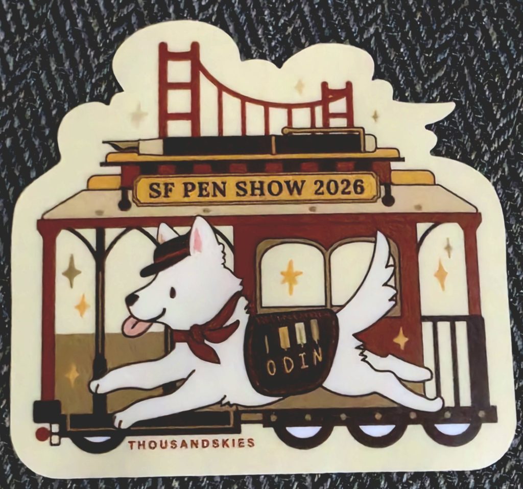 The SF Pen Show 2026 show sticker, depicting a red cable car with a large fountain pen on top, and above that a depiction of the Golden Gate Bridge. In front of the cable car is a white dog, with a coat that says "ODIN".